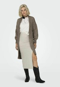 Cárdigan de punto marrón con mangas de gran tamaño, superpuesto a una camiseta blanca y una falda gris con una abertura lateral, combinado con botas de cowboy negras.