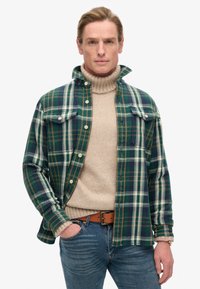 Camisa de franela a cuadros en verde y azul marino con dos bolsillos en el pecho, llevada sobre un suéter de cuello alto beige, combinada con jeans azules y un cinturón marrón.