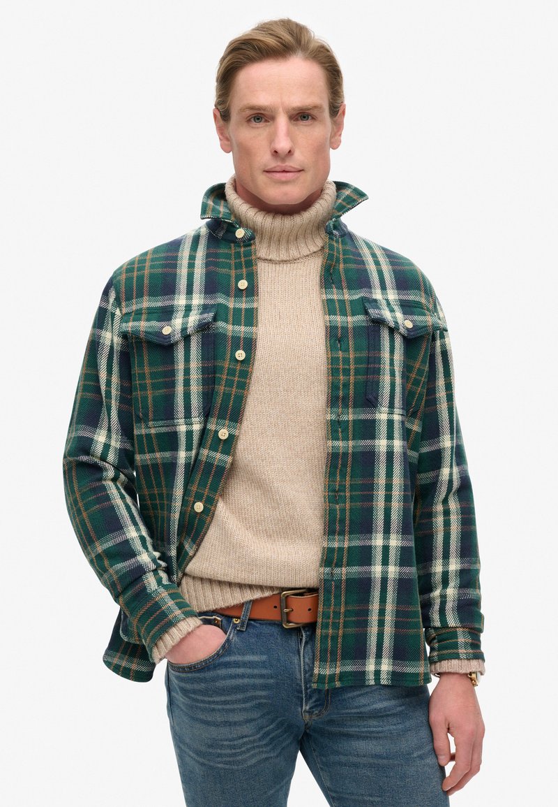 Camisa de franela a cuadros en verde y azul marino con dos bolsillos en el pecho, llevada sobre un suéter de cuello alto beige, combinada con jeans azules y un cinturón marrón.