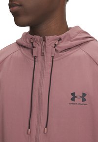 Rožinė lengva medžiaga užtrauktuku užsegama striukė, kuri turi gobtuvą, priekinį užtrauktuką ir juodą "Under Armour" logotipą. Aiškus siuvimas ir reguliuojamos raišteliai.