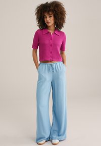 Rozā adīta īsām piedurknēm krekls ar pogu aizdari, saskaņots ar gaiši ziliem platām biksēm ar elastīgu jostasvietu.