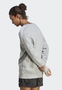 Siv sv sweatshirt s tremi belimi črtami vzdolž ramen, rahlo prevelik kroj, rebrast rokav in rob, oblečen nad črnimi kratkimi hlačami.