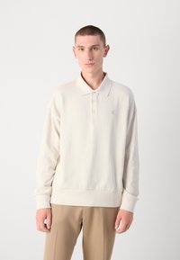 Cremefarbenes Poloshirt mit Kragen, dreiknöpfiger Knopfleiste, gerippter Textur und einem dezenten Logo auf der Brust. Weicher Stoff, lässige Passform.