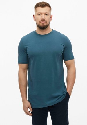 Bärtiger Mann mit kurzen Haaren, der ein türkisfarbenes T-Shirt mit kurzen Ärmeln und dunkle Hosen trägt, steht mit einer Hand in der Tasche vor weißem Hintergrund.