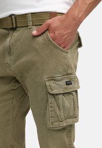 Uomo che indossa pantaloni cargo verde oliva con mano nella tasca frontale, cintura di tessuto visibile e tasca laterale con patta.