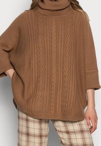 Femme portant un pull à col roulé marron en tricot torsadé avec des manches trois-quarts et un pantalon beige à carreaux, main dans la poche, sur un fond clair.