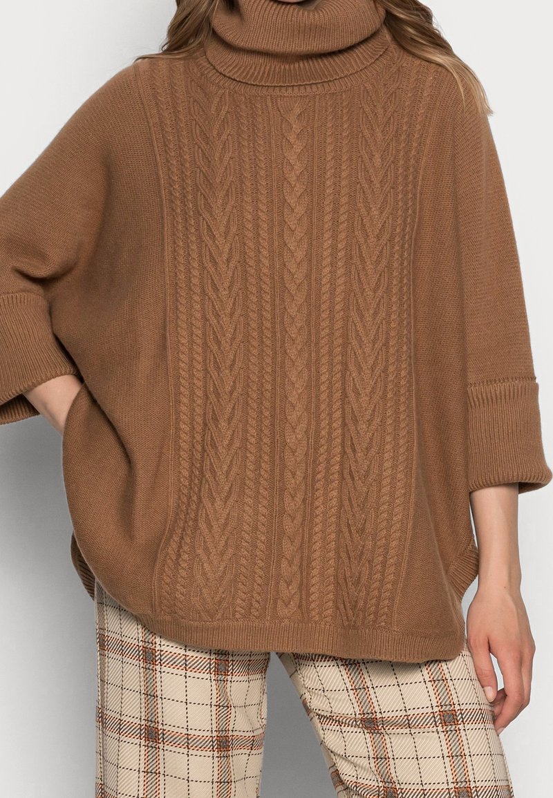 Femme portant un pull à col roulé marron en tricot torsadé avec des manches trois-quarts et un pantalon beige à carreaux, main dans la poche, sur un fond clair.