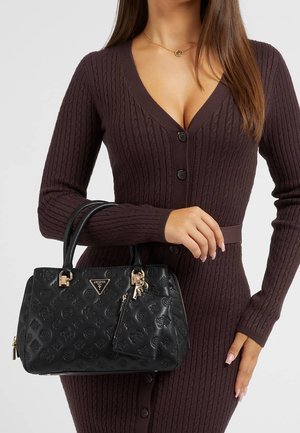 Woman wearing a brown ribbed button-up dress holding a black embossed Guess handbag with gold accents and a matching pouch.