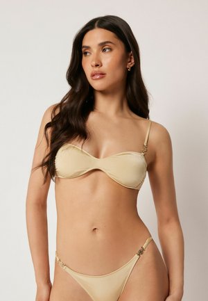 Femme aux longs cheveux foncés portant un bikini deux pièces beige, debout devant un fond clair uni, regardant vers sa gauche.