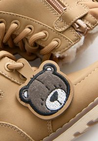 Braune Winterstiefel mit braunen Schnürsenkeln, einem Reißverschluss, Wollfutter und einem daran befestigten Teddy-Gesichtsschild.