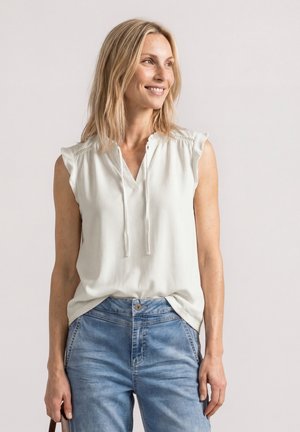 Femme souriante aux cheveux blonds portant un chemisier blanc sans manches et un jean bleu, debout devant un fond clair uni.