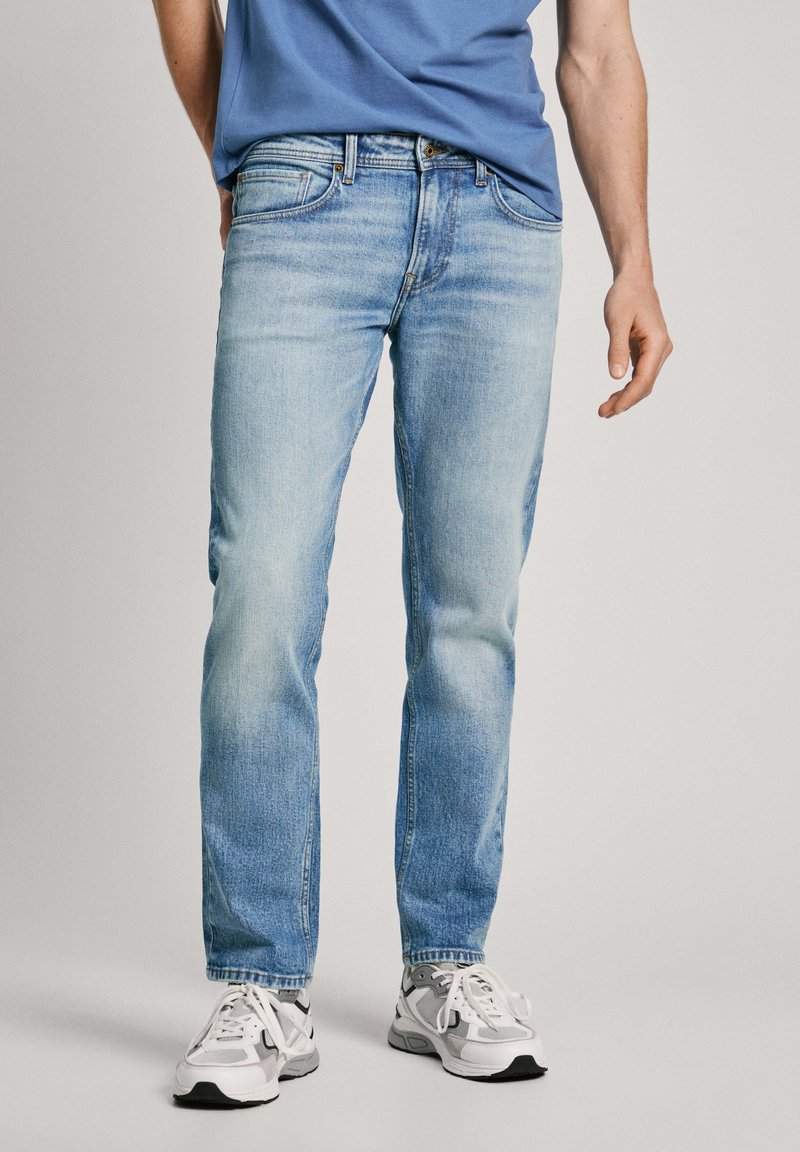 Vaqueros de mezclilla azul claro con corte recto, acabado desgastado, diseño de cinco bolsillos y costuras en contraste, combinados con zapatillas blancas y grises.
