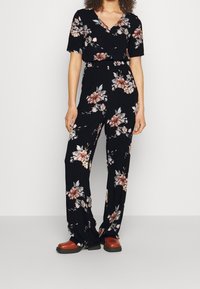 Kobieta ubrana w czarny kombinezon w kwiaty z krótkimi rękawami i szerokimi nogawkami, w połączeniu z brązowym obuwiem, stojąca na tle jednolitym.