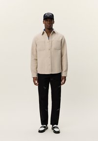 Jachetă din fleece bej deschis, cu două buzunare pe piept, purtată peste o cămașă cu model. Asortată cu pantaloni negri deteriorați și pantofi alb-negru.