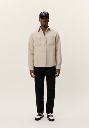 Light beige fleece shirt jacket with two chest pockets, worn over a patterned shirt. Paired with distressed black pants and black and white shoes.