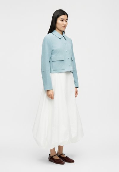 Γυναίκα που στέκεται φορώντας ένα ανοιχτό γαλάζιο κοντό μπουφάν με μεγάλα κουμπιά, λευκή αέρινη φούστα και μπορντό παπούτσια Mary Jane, πάνω σε απλό φόντο.