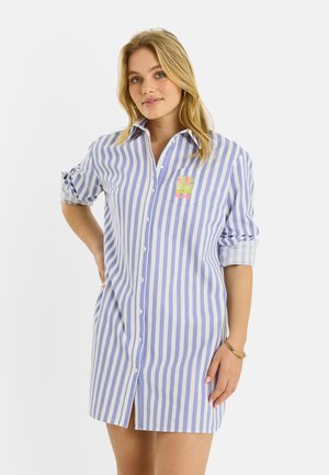 Femme blonde portant une robe-chemise à boutons à rayures verticales bleu et blanc, manches retroussées, avec un petit écusson floral coloré.