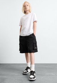Hvid kortærmet t-shirt, sorte cargo-shorts med lommer og sorte sneakers med hvide detaljer. Modellen står med hænderne i shorts' lommer.