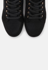 Black sneakers with textured fabric and suede materials. Features round black laces and gold-tone eyelets. Rounded shoe toe design.