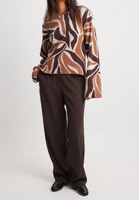 Stickad tröja i brun och grädde abstrakt mönster, långa ärmar, rund halsringning, i kombination med lösa mörkbruna byxor och glänsande svarta skor.