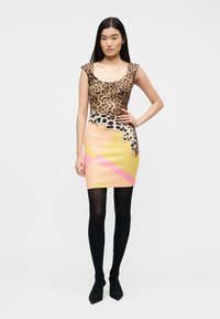 Γυναίκα με αμάνικο μίνι φόρεμα με τοπ λεοπάρ εκτύπωσης και φούστα με γεωμετρικά μοτίβα σε παστέλ κίτρινο, ροζ και πορτοκαλί, μαύρες καλτσοδέτες και μαύρα τακούνια.