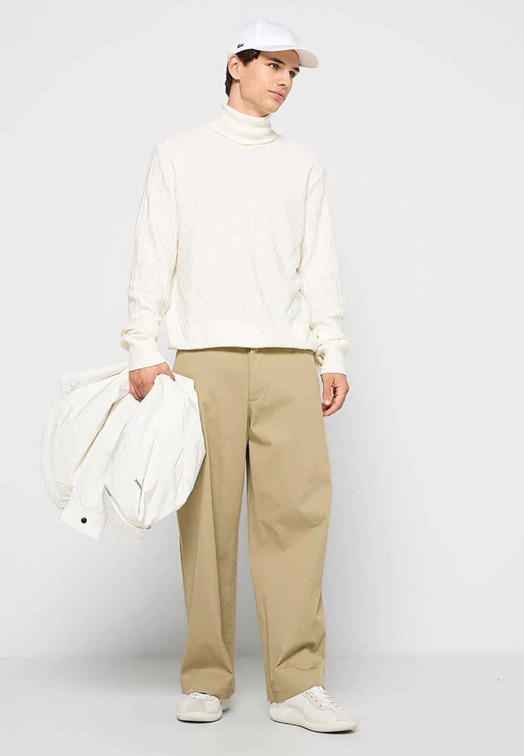 Maglione bianco a costine con collo alto, pantaloni beige a gamba larga, sneakers bianchi e berretto bianco. Una giacca bomber bianca è tenuta nella mano sinistra.