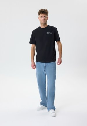 Hombre de pie con camiseta negra con estampado gráfico, jeans azul claro y zapatillas blancas sobre un fondo claro y liso.