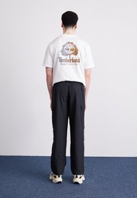 Hvid t-shirt med et brunt og blåt Timberland-logo på ryggen, parret med sorte cargo-bukser og lyse sneakers.