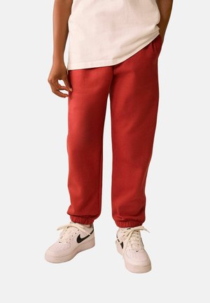 Person iført røde joggingbukser og hvide Nike-sneakers med sort swoosh, står med den ene hånd i lommen mod en ensfarvet baggrund.