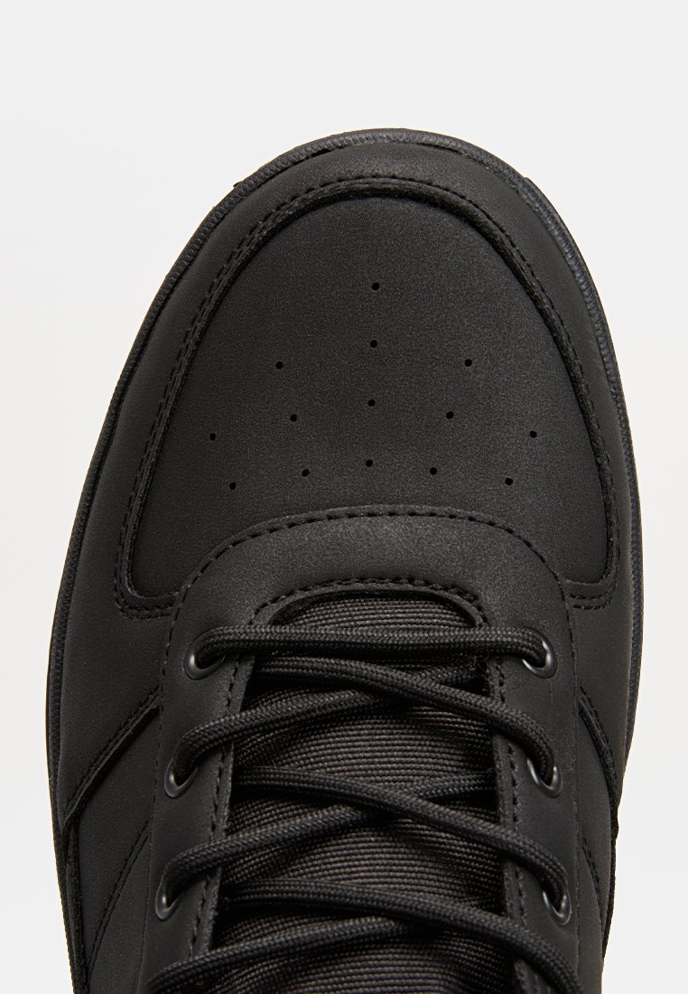 Scarpa sportiva nera con tomaia sintetica liscia, punta perforata, lacci rotondi e suola resistente. Presenta dettagli cuciti e fori per lacci.