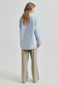 Cămașă din bumbac de culoare albastru deschis, cu tiv curbat, slituri laterale și mâneci lungi, asortată cu pantaloni largi cu dungi bej.