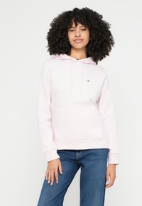 Jasnoróżowa bluza z kapturem, z przednią kieszenią, sznurkiem przy kapturze oraz małym logo na lewej piersi. Miękki materiał i luźny krój, zestawiona z niebieskimi dżinsami.