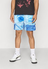 Lyseblå mesh-shorts med geometriske mønstre i varierende blånyanser. Har en solid blå applikasjon på det ene benet. Brukes med hvite sokker og sko.