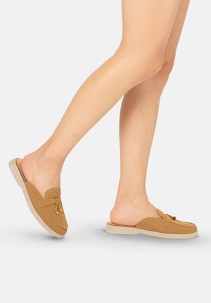 Piernas con mocasines de ante color beige con borlas decorativas sobre un fondo gris claro, un pie ligeramente levantado.