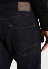 Jeans in denim blu scuro con cuciture pronunciate, una tasca posteriore con un design a forma di V e un'etichetta del marchio color cammello sopra la tasca destra.