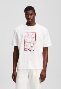 Balta medvilninė marškinėliai su išskirtiniu priekinio dizaino piešiniu, raudonais akcentais ir juodu script stiliaus užrašu "Karl Lagerfeld".