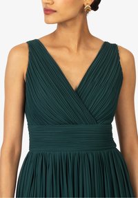 Grön plisserad klänning med djup V-ringning och bred midjeband. Har mjuk textur och rynkor längs midjan som framhäver silhuetten.