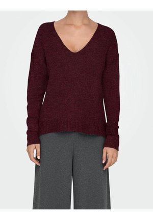 Woman wearing a burgundy V-neck sweater with long sleeves paired with loose-fitting dark gray pants, standing against a plain background.