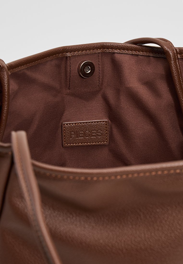 Interieur van een bruine leren tote bag met een gladde textuur, voorzien van een drukknoopsluiting en een etiket met de opdruk "PIECES."