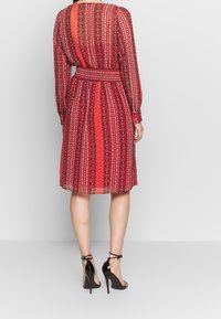 Robe rouge à motifs avec des manches longues, taille cintrée et ourlet au niveau des genoux. Présente un mélange de designs géométriques et floraux. Sandales à talons hauts noires.