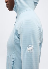 Άτομο που φοράει ανοιχτό μπλε φούτερ με κουκούλα και λογότυπο Mammut στο πάνω μέρος του μανικιού, εμφανιζόμενο σε προφίλ μπροστά σε απλό φόντο.