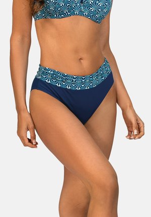 Kobieta nosząca niebieskie, wysokie figi bikini z wzorzystym paskiem i dopasowany top bikini z tym samym wzorem, stojąca na jednolitym tle.