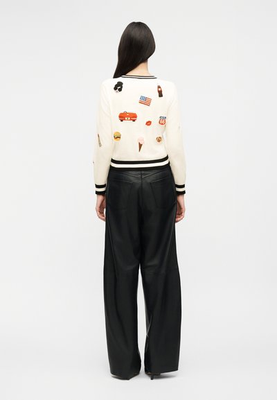 Γυναίκα που φοράει λευκό πουλόβερ με πολύχρωμα μπαλώματα αμερικανικού θέματος και μαύρο δερμάτινο παντελόνι με φαρδιά μπατζάκια, στρέφεται προς τα πίσω σε απλό φόντο.