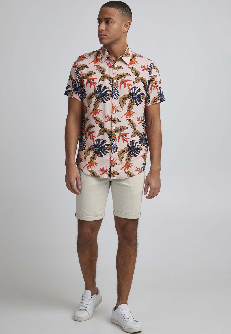 Kortermet skjorte i lyserødt stoff med tropiske bladmønstre i marineblått, grønt og oransje. Brukes med beige rullede shorts og hvite joggesko.