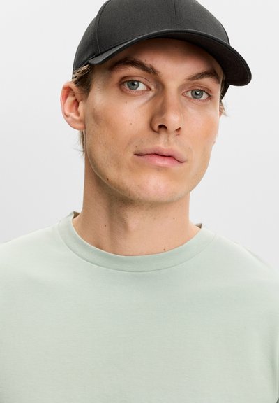 Giovane uomo con pelle chiara, occhi azzurri, che indossa un cappello nero e una maglietta con scollo tondo verde chiaro, che guarda leggermente di lato.