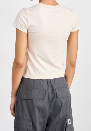 Personne portant une chemise beige unie à manches courtes et un pantalon cargo gris foncé, vue de dos sur un fond uni.