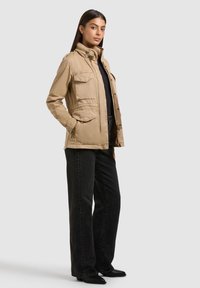 Donna che indossa una giacca utility beige sopra una maglietta nera, con jeans neri a gamba larga e scarpe nere con punta appuntita, in piedi davanti a uno sfondo semplice.