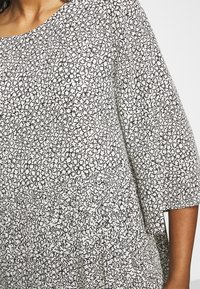 Mustavalkoinen kuvioitu paita, jossa on pyöreä pääntie, kolmanneksen pituiset hihat ja rypytetty kohta vyötäröllä, sekä kukkakuviot.