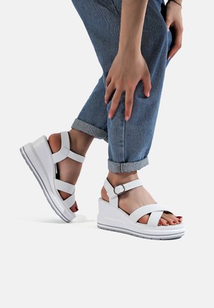Sandali wedge bianchi con doppie cinture alla caviglia, design a cinturino incrociato e suola testurizzata. Materiale sintetico liscio, tacco impilato e fibbia regolabile.