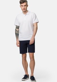 Biała bawełniana koszulka polo z kołnierzykiem, w zestawieniu z granatowymi szortami. Model ma na sobie niebieskie trampki, prezentując tatuaż na lewym ramieniu.
